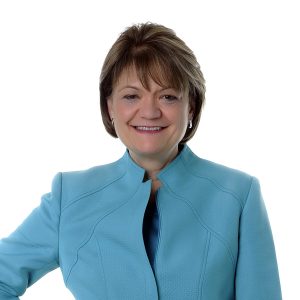 A professional looking individual with short hair is wearing a light blue blazer and smiling at the camera against a white background.