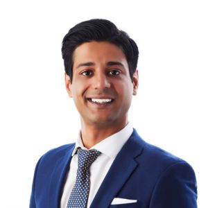 A man in a blue suit and polka-dot tie is smiling at the camera with a plain white background.