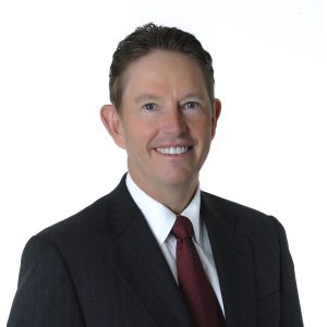 A man in a dark suit and red tie is smiling against a white background.