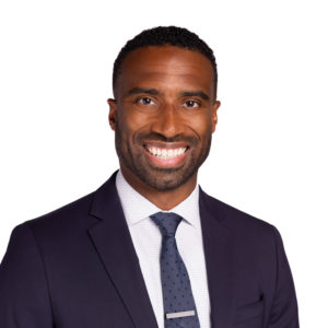 A man is smiling while wearing a dark suit, shirt, and tie against a white background.