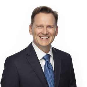 A man in a dark suit and blue tie is smiling against a white background.