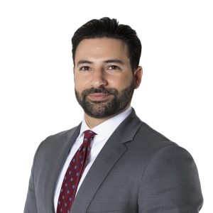 A well-dressed individual with a beard is smiling and posing against a white background.