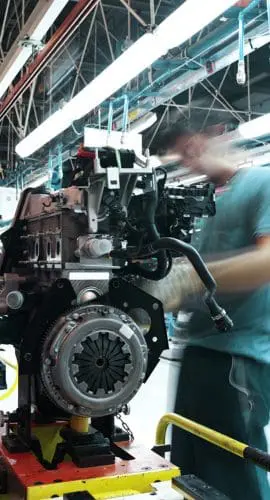 A worker assembles car engines on a factory production line with industrial equipment and fluorescent lighting overhead.
