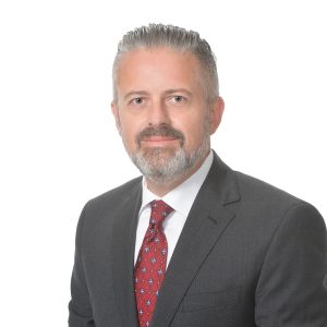 A professionally dressed individual with a beard and mustache is posing for a portrait with a white background.