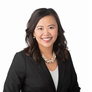 A person with dark hair, wearing a black blazer and a pearl necklace, is smiling against a white background.