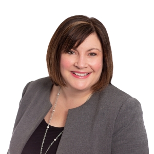 A smiling individual with short hair is wearing a grey blazer over a dark top and a silver necklace.