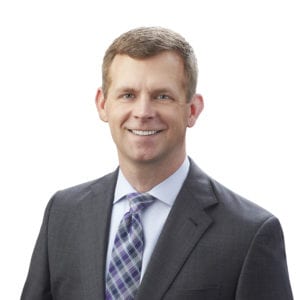 A person is dressed in a gray suit, white shirt, and patterned tie, smiling against a plain white background.