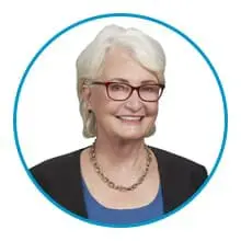 A person with white hair, wearing a black blazer, blue top, red glasses, and a necklace, is smiling within a circular border.