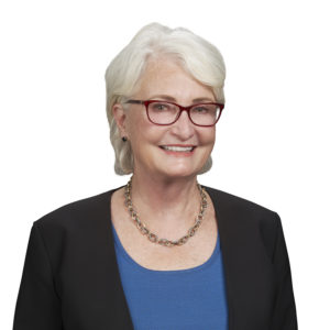 An individual with short white hair, red glasses, and a necklace is smiling while wearing a blue top and a black jacket.