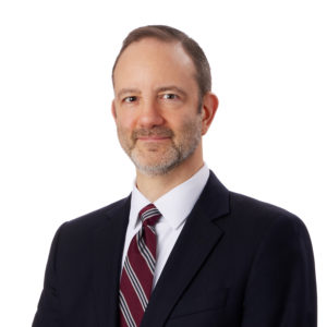 A man is dressed in a dark suit with a maroon, white, and gray striped tie against a plain white background.