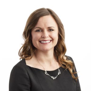A woman with long brown hair is smiling while wearing a black top and a necklace.