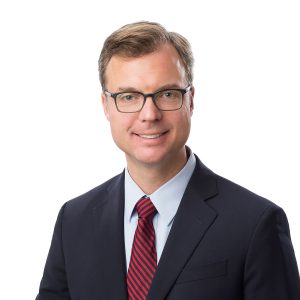 A person with glasses is wearing a suit with a red tie against a white background.