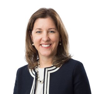 A professional-looking individual with shoulder-length hair, wearing a necklace and a striped blazer over a white top, is smiling at the camera.