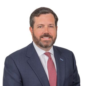 A man with short hair and a beard is wearing a suit with a white shirt and a pink tie, smiling in front of a plain white background.