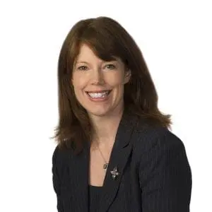 A person is smiling while wearing a dark blazer and a necklace, set against a plain white background.