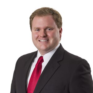 A smiling individual wearing a dark suit with a white shirt and red tie poses against a plain background.