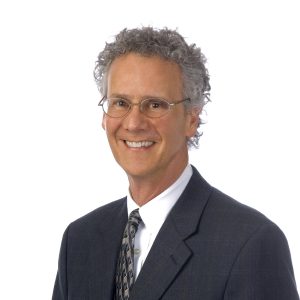 A smiling individual with curly gray hair is wearing a business suit and tie against a white background.