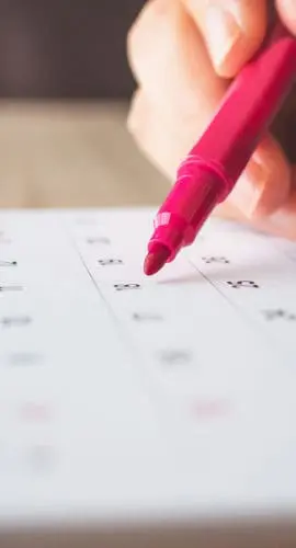 A hand is using a red pen to mark a date on a calendar.
