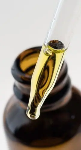 A droplet of yellow liquid is suspended from a dropper above an open brown bottle.