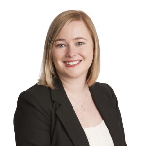 A smiling person with blonde hair wearing a black blazer and a white top against a white background.