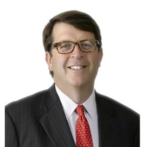 A smiling individual wearing glasses, a dark suit, and a red patterned tie against a light background.