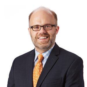 A smiling person with glasses wearing a business suit, a patterned blue shirt, and an orange tie.