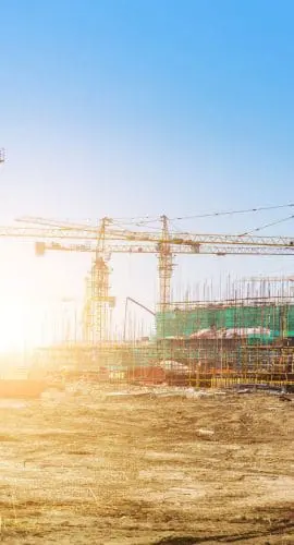 A construction site with multiple cranes and a scaffolding structure illuminated by bright sunlight.