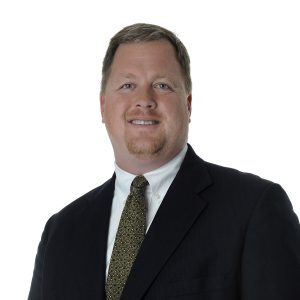 A smiling individual wearing a dark suit, white shirt, and patterned tie against a white background.
