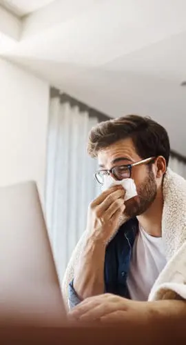 A person wrapped in a blanket works on a laptop while holding a tissue to their nose.