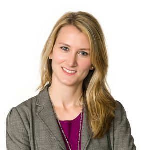 A smiling woman with long blonde hair wearing a gray blazer and a purple blouse.