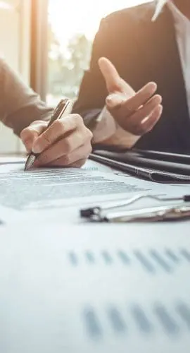 A person is signing a document next to another individual gesturing with their hand, with paperwork and electronic devices on the table in a brightly lit office setting.