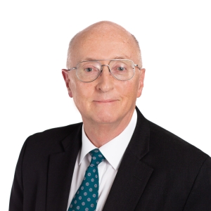 A balding individual with glasses wearing a black suit, white shirt, and patterned tie is posing against a white background.