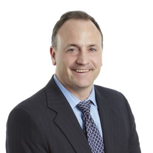 A smiling person wearing a professional suit with a blue tie and a patterned shirt.