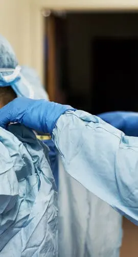 Two medical professionals wearing protective gear are assisting each other with their gowns.