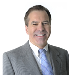 A smiling individual wearing a gray suit with a light blue tie and white shirt against a white background.
