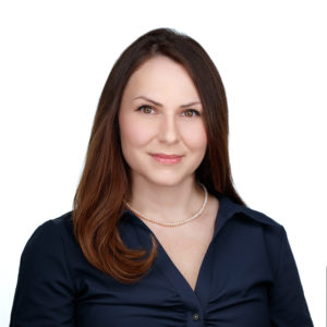 A person with long brown hair is wearing a dark blue blouse and a pearl necklace, smiling against a white background.