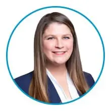 A person with long, straight hair is wearing a dark blazer over a white shirt and is smiling at the camera, enclosed within a blue circular border.