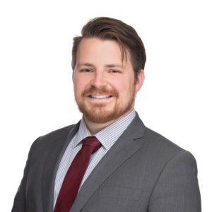 A person with short, brown hair and a beard is wearing a gray suit, a white shirt with gray stripes, and a red tie, smiling against a white background.