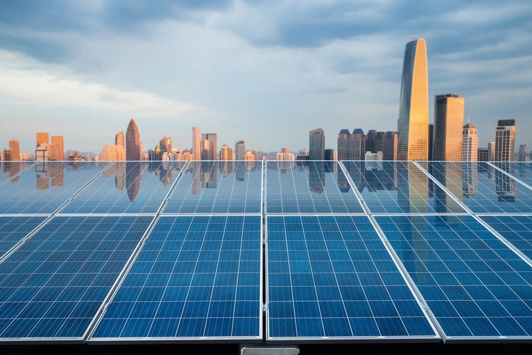 An array of blue solar panels is in the foreground with a city skyline under a cloudy sky in the background.