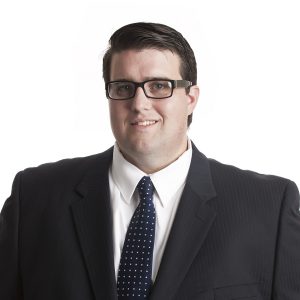 A man wearing glasses, a black suit, white shirt, and a blue tie with polka dots is posing for a professional portrait against a plain background.