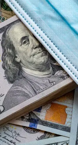 A stack of hundred-dollar bills is partially covered by a blue surgical mask.