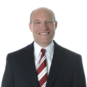 A smiling individual with a bald head wearing formal attire including a red tie and a white shirt against a white background.