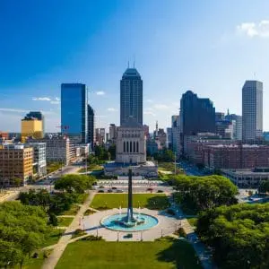 Aerial view of a city skyline with a park and a prominent fountain in the foreground under a clear blue sky.