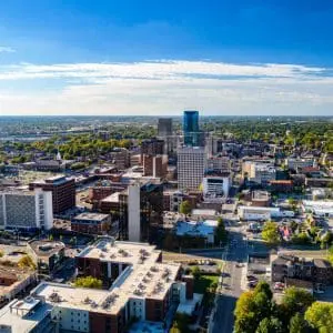 An aerial view of a downtown district showcases a mix of high-rise buildings, urban streets, and scattered greenery under a blue sky with light cloud cover.