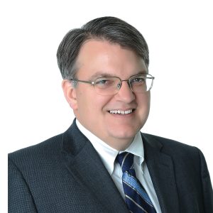 A smiling individual with glasses is wearing a dark suit and a blue tie with a pattern, against a light background.