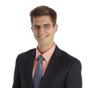 A smiling person wearing glasses, a black suit, a striped tie, and a coral-colored shirt against a white background.