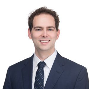 A person wearing a dark suit, white shirt, and a dark polka-dot tie smiles against a plain white background.
