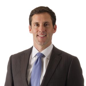 A person is wearing a dark suit, a light blue tie, and a white shirt, smiling against a plain background.