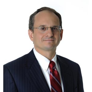 A person wearing glasses, a dark suit, a white shirt, and a red striped tie poses against a white background.