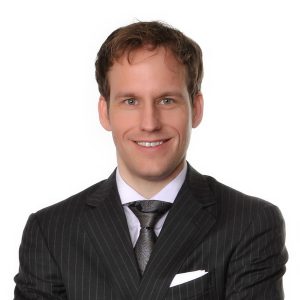 A smiling individual with short hair is wearing a pinstripe suit and tie.